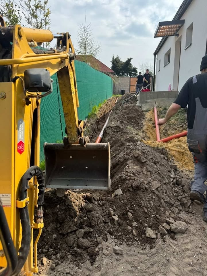Yellow excavator with open bucket digging soil at construction site next to green fence and white building
