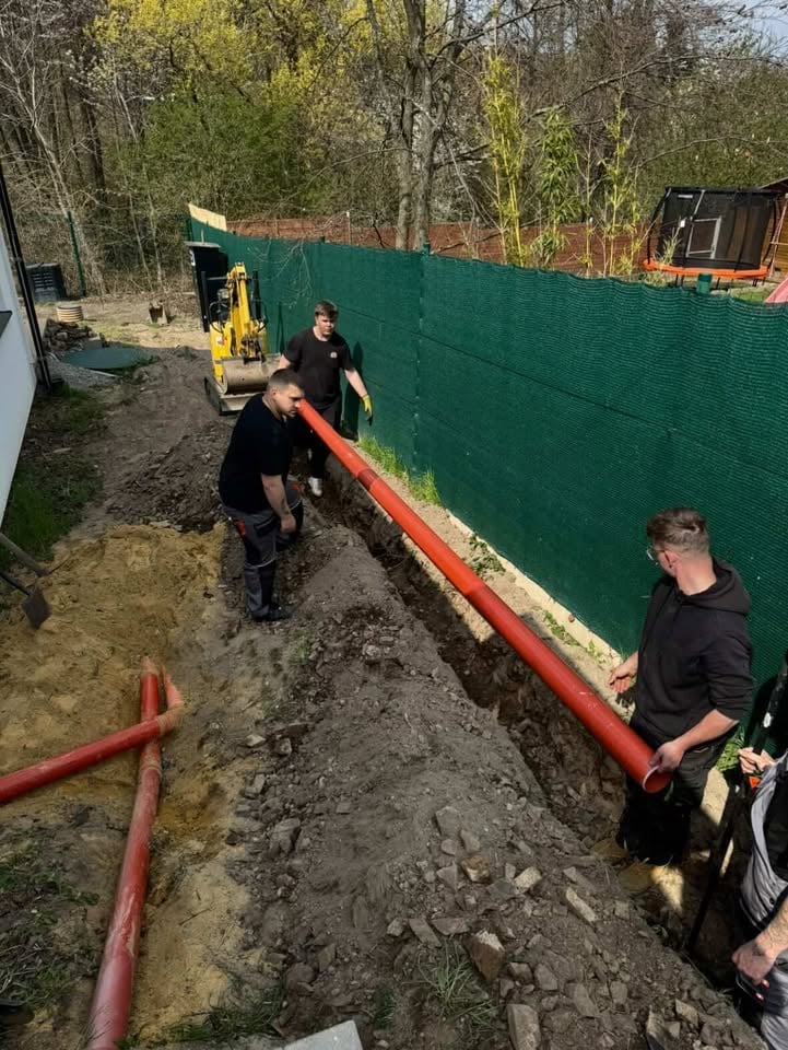 Three workers installing a green fence with a red base along a dirt path surrounded by bare trees and equipment