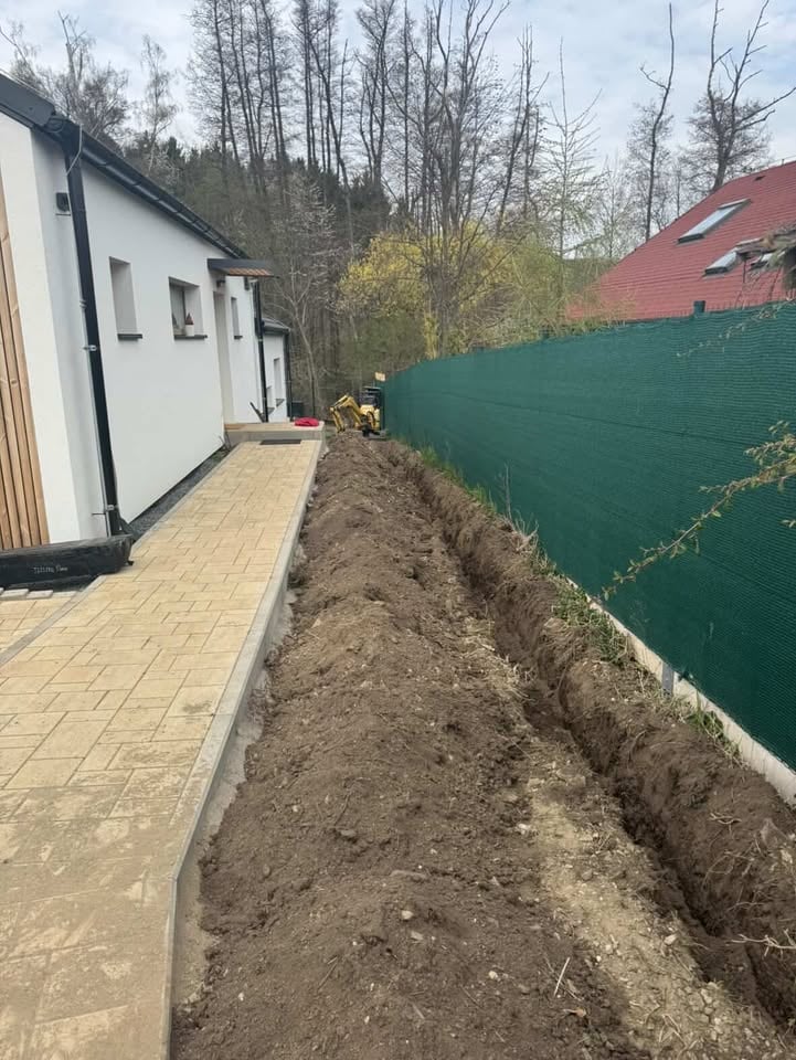 Narrow dirt trench beside a white cottage with green fence and bare trees in background